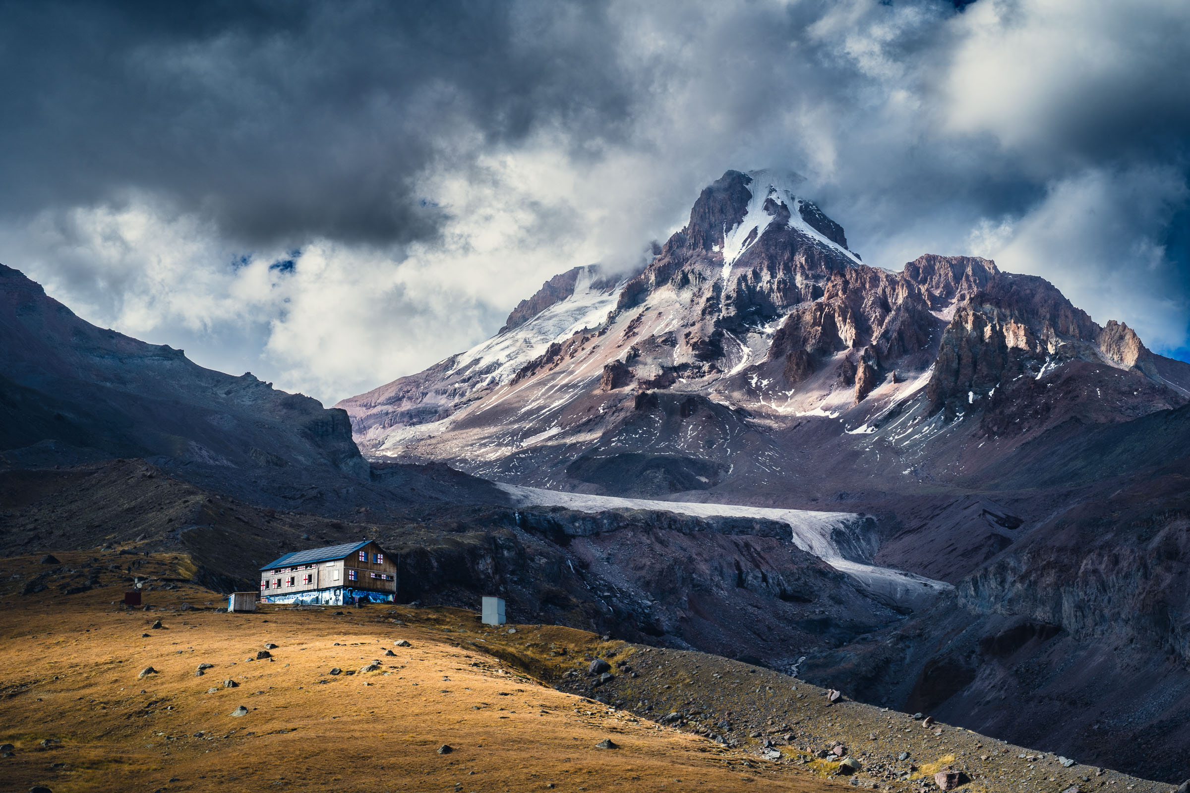 A small mountain hut stands on a sunlit golden ridge below the snow-capped summit of Mount Kazbek and the Gergeti Glacier, under dramatic clouds.