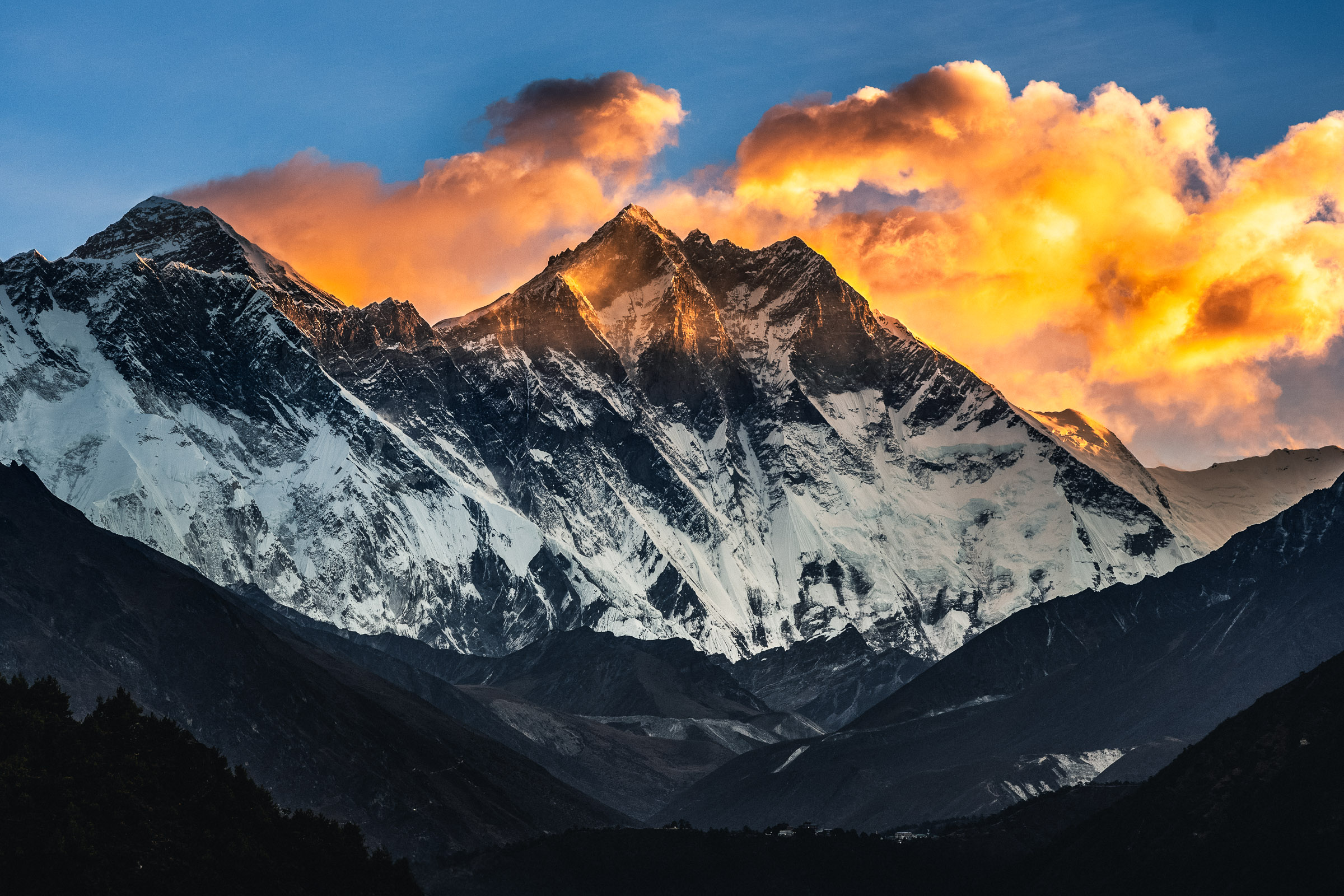 Orange-gold clouds rise behind the snow-covered wall of Mount Everest and Lhotse, above dark valley slopes beneath a blue sky.