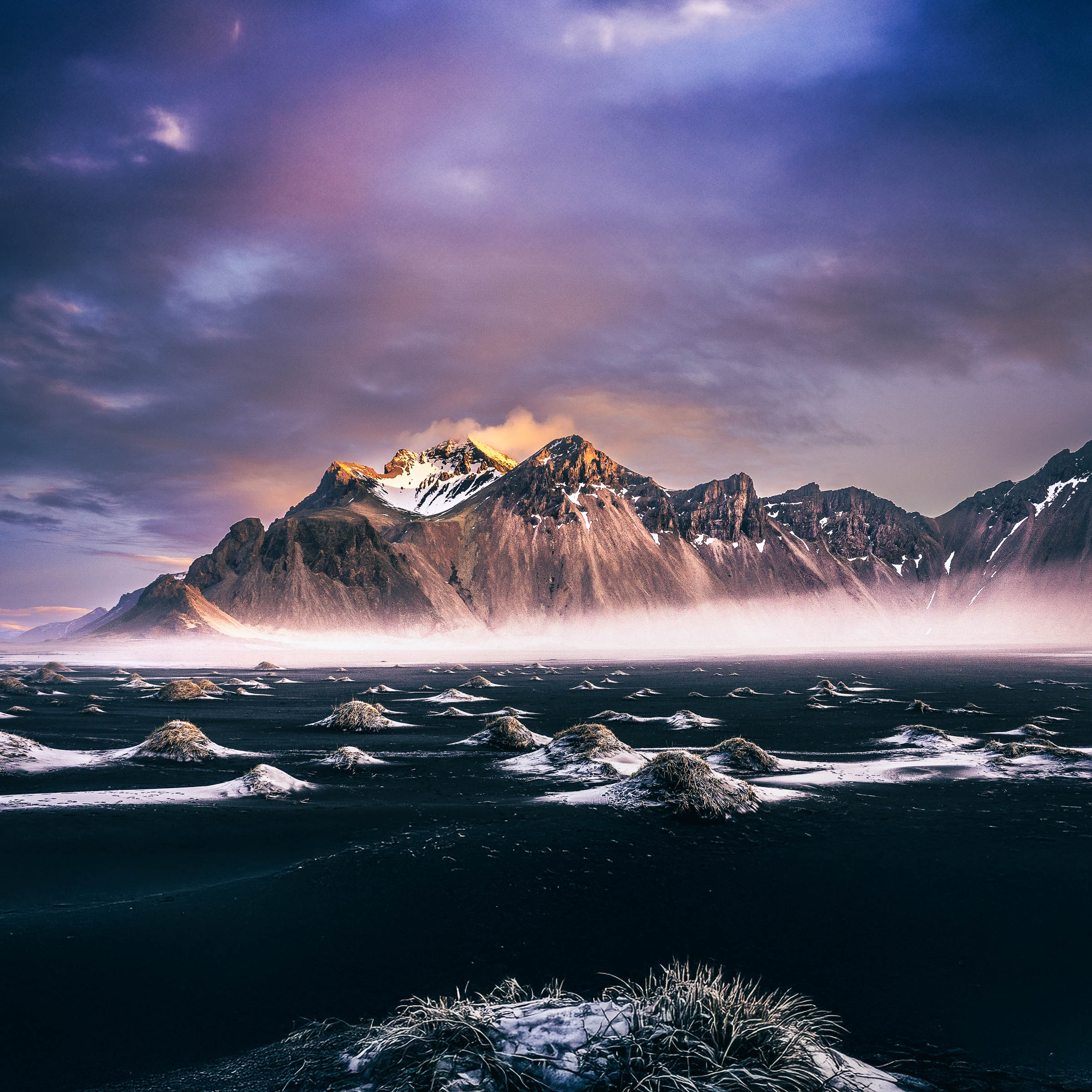 A sunlit snowy summit of Vestrahorn rises above low mist and black sand dunes with frosted grass tufts, beneath a purple cloudy sky.